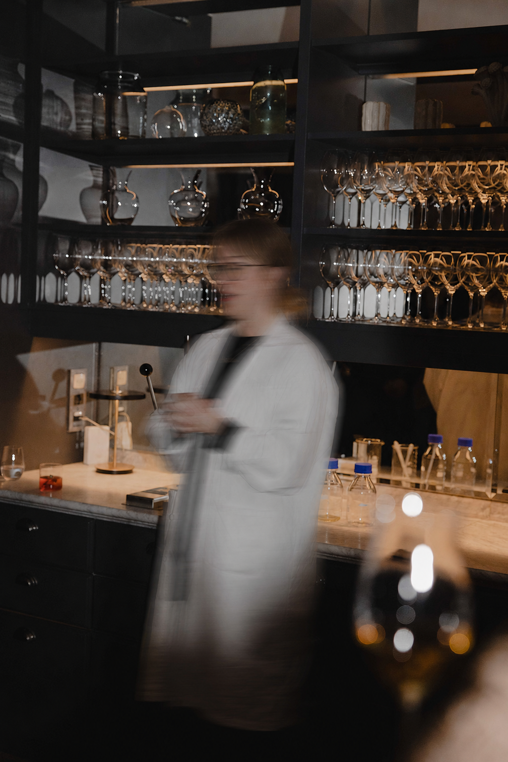Perfumer in a white uniform stands blurred in a dimly lit bar with shelves of glassware and bottles in the background.