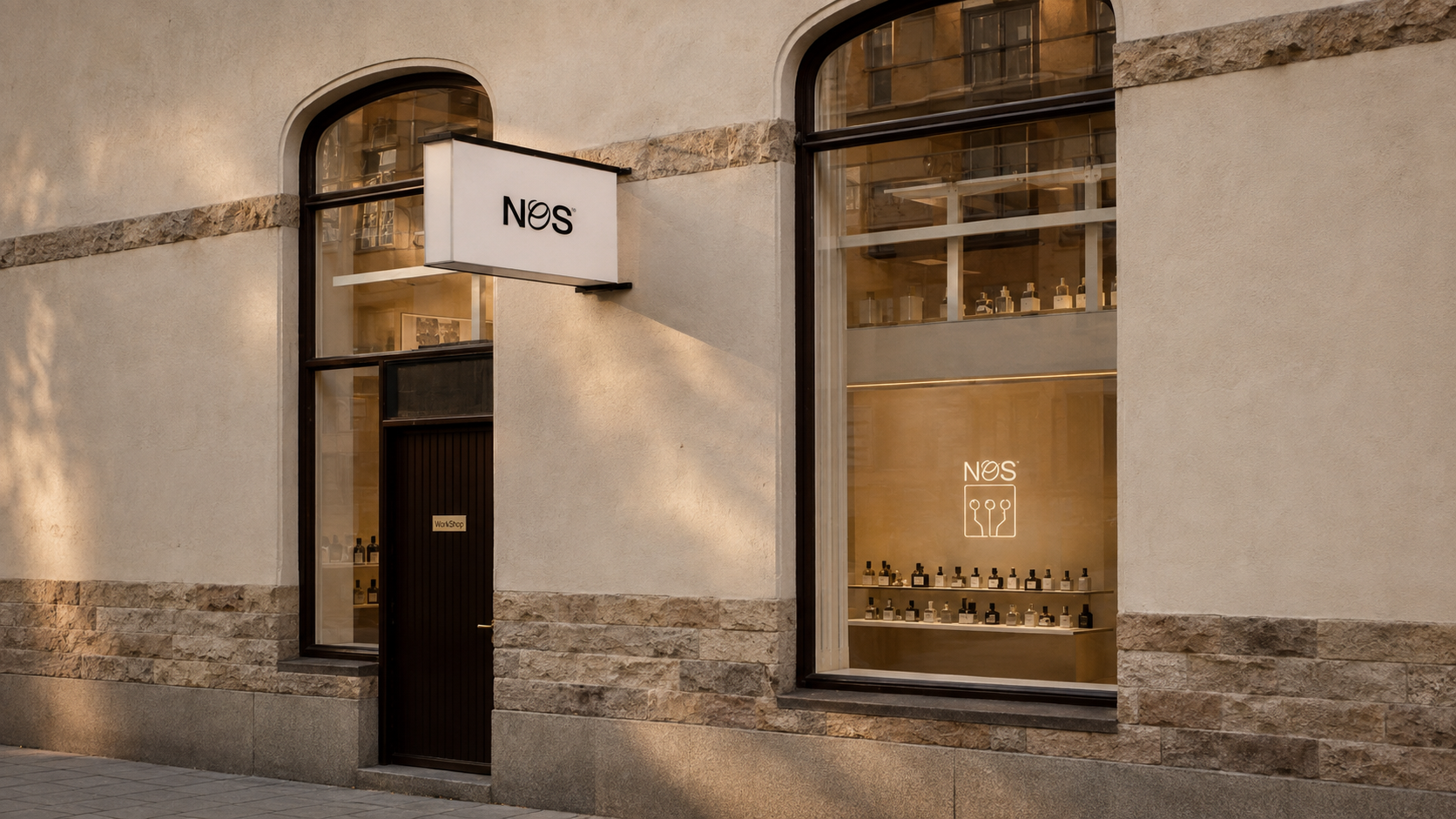 A storefront exterior with a beige stucco wall, stone trim, a dark door, and large arched windows. A white hanging sign reads NOS, and the window display also shows the NOS logo and bottles.