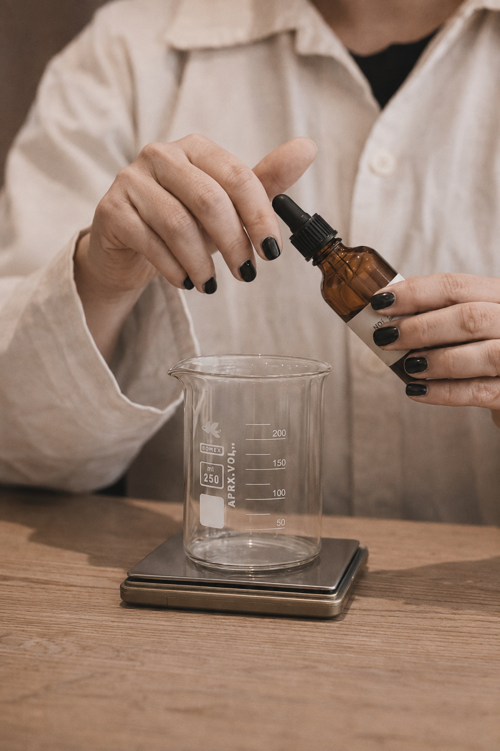 A person in a light beige lab coat holds an amber dropper bottle over a clear glass beaker on a digital scale, with a wooden tabletop in the background.
