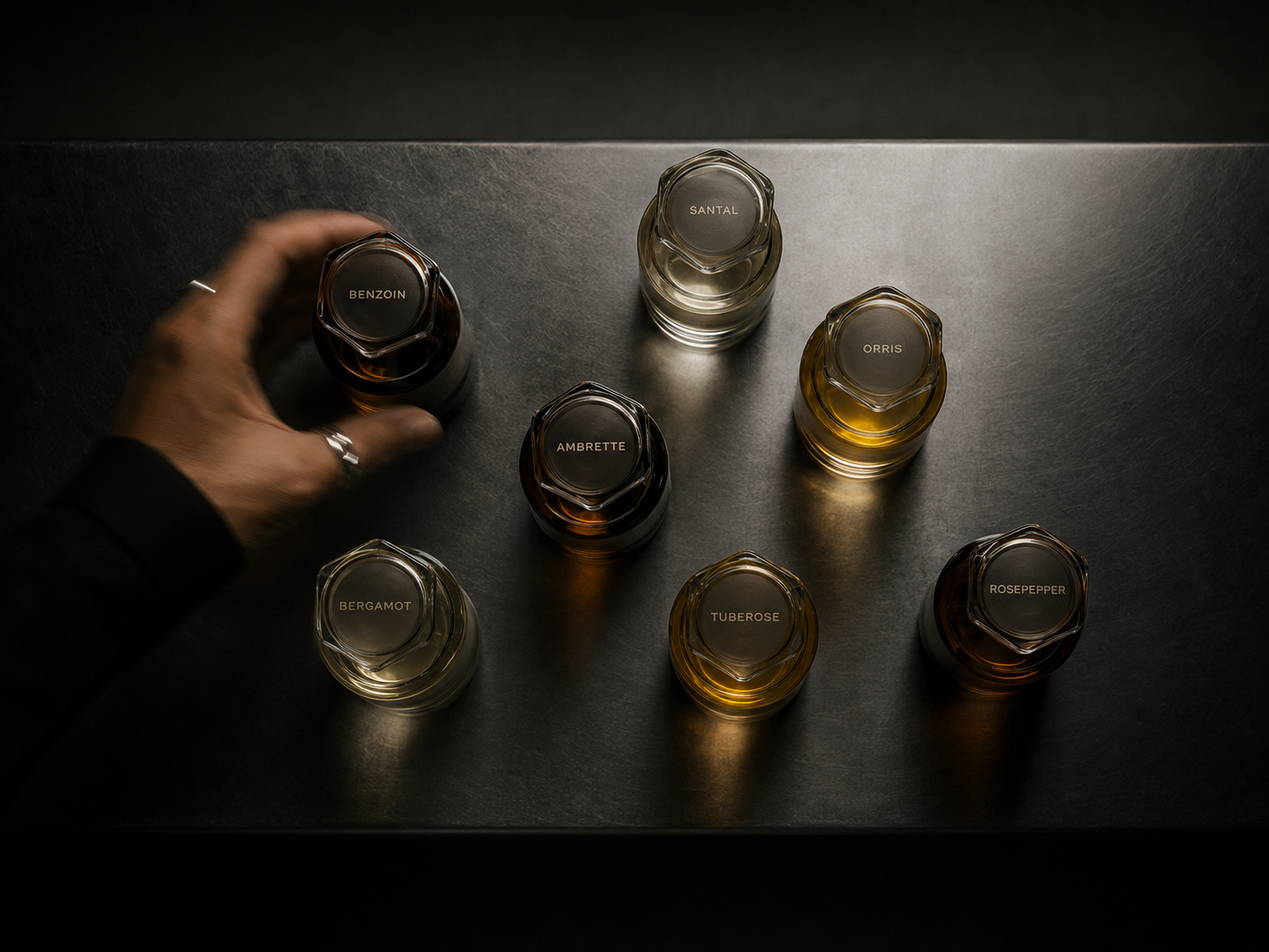 A hand reaches toward a row of faceted glass perfume bottles on a dark tabletop. The bottles contain amber, gold, and clear liquids, with silver-toned caps labeled with fragrance names such as Bosphore, Santal, Saffron, Ambrette, Turquoise, and Bosphore.